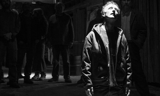 Actor Paul Cram on his knees looking up to a bright light, in a black and white image from the set of the film "Anniversary"